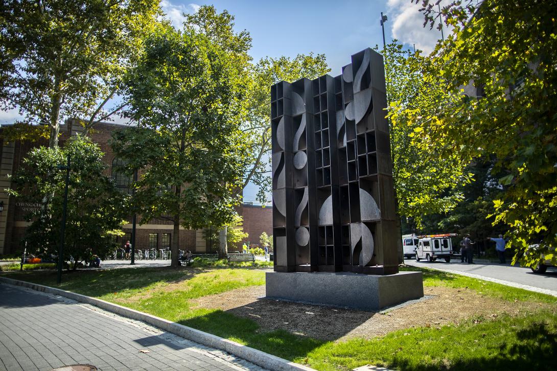 Large abstract steel sculpture with trees and campus buildings in background 